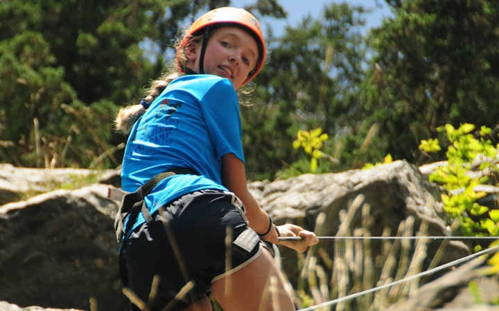 A young person wearing safety gear is secured by ropes as they look down toward the camera while rock climbing. 
